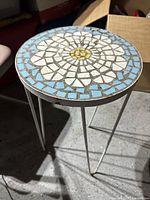 Round end table with intricate mosaic tile top in blue, white, and yellow on metal legs.