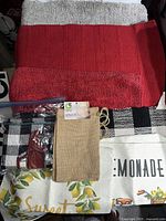 Lot displaying red and beige bath mats, checkered tablecloth, lemon-themed cloths, and burlap pouch laid out on a surface.