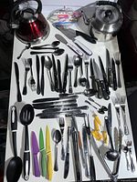 Overview of the kitchen utensils laid out on a white surface showing teapots, knives, forks, spoons, ladles, and cooking utensils