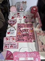 Photo showing various Valentine’s Day decorative signs including wooden gnome holding 'Be Mine' heart, heart-shaped and rectangular signs, and a pink 'Kissing Booth' sign.