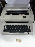 Front angle photo of IBM Selectric typewriter with beige body and black keys, showing keyboard and upper carriage area. Small IBM card placed in front.