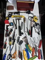 Wide view of all assorted hand tools and cutting tools laid out including pliers, hammers, screwdrivers, Allen keys, knives, scissors, measuring tapes, and a red metal toolbox