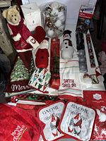 Photo of assorted large holiday fabric decorations and plush figures arranged together, including mats, mitts, towels, stuffed Santa and snowman, and ornaments in clear box.