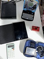 Overview of untested electronics on a white table including iPad, DVD player, Batman handheld game, Marlboro camera, calculator, and portable GPS device.