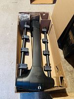 Top-down view of the bike rack in its original box showing the main support bar and two side arms with attached rubber clamps. Plastic mounting hardware is visible in a separate compartment.