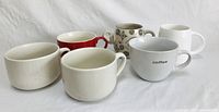 Six ceramic coffee mugs in different colors and designs arranged in two rows on white fabric background. Included red, off-white, white with text, and patterned mugs.