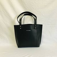Front view of black leather tote bag on white background showing full tote shape and dual handles.