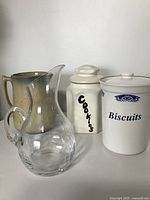 Front view of two ceramic cookie jars labeled 'Cookies' and 'Biscuits', one beige ceramic jug, and clear Vilca glass pitcher.
