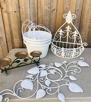 Items displayed on concrete floor against wooden fence showing white metal wall hangings, hanging planter basket, white wire planter bucket, and green metal candle holder.