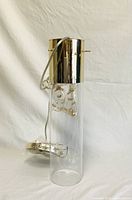 Light fixture shown upright with clear glass cylinder and attached gold-colored metal top. Decorative interior amber crystals and wire visible inside. Power cord included and coiled inside glass.