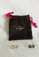 Two pairs of Kate Spade stud earrings and a black dust bag with pink drawstrings featuring the Kate Spade logo in gold text.