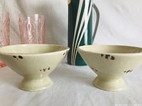 Two beige ceramic footed bowls with crackled glaze and dark spots, several glasses, and a green and white striped jug visible.