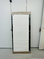 Large rectangular mirror wrapped in plastic and cardboard for protection, standing upright against a white wall.