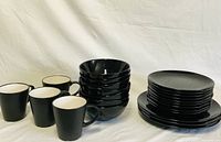 Photo of black ceramic bowls stacked next to stacks of large and smaller plates and four black coffee cups with white interiors.