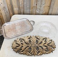 Photo of three trays on table: rectangular silver tray with handles, clear round glass tray, and oval carved wooden tray