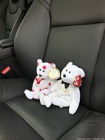 Two small Beanie Babies seated on a car seat, white bear with red spots and solid white bear with a necklace and TY tag visible