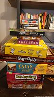 Photo showing stacked board and card games including Diary of a Wimpy Kid Cheese Touch, Catan Junior, Pug Opoly, Say Anything, Pass the Popcorn, Sushi Go!, Quick Draw Charades, and Card Games box. Minecraft game visible on top side.