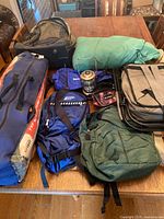Camping gear on wooden table showing two tents in carry bags, various bags, camping lantern and tent poles.