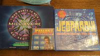 Photo shows Who Wants to Be a Millionaire board game box, Jeopardy computer game CD case with Alex Trebek image, and sealed Jeopardy board game box.