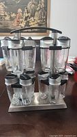 View of the glass spice rack showing 12 glass spice bottles in a metal stand with handle, bottles have black plastic lids and dispensing tops.