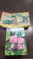 Two unopened LEGO sets placed on a dark surface, boxes show noticeable damage and creasing. Top box is LEGO Creator 3-in-1 dragon build, bottom box is LEGO Minecraft pig with baby zombie.