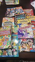 Photo showing eleven used Pokémon books of various sizes and titles laid out on a table, featuring colorful covers with Pokémon characters and titles.