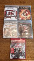 Five PS3 game cases on wooden table showing front covers: Dragon Age Origins Ultimate Edition, Heavenly Sword, God of War Origins Collection sealed, Darkness II, Dead Island GOTY Edition.
