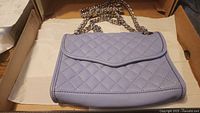 Front view of light purple quilted leather purse with flap closure and chain strap inside a box.