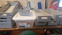 Front view of various gray plastic storage containers arranged on a table, different sizes and types visible including one with a lid and a small woven basket