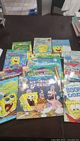 Photo showing a collection of more than 10 Spongebob Squarepants books arranged on a table. Books vary in size and feature colorful cartoon covers depicting Spongebob and friends.