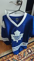 Blue Toronto Maple Leafs hockey jersey with white collar and front logo shown on hanger.