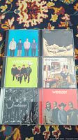 Top view of 6 Weezer CD jewel cases laid out showing front album covers with band photos and artwork.