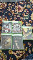 Lot of five Xbox 360 action game cases laid out on patterned carpet, including Splinter Cell Conviction, Sleeping Dogs, The Saboteur, Condemned 2: Bloodshot, and Dead Space 2.