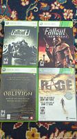 Four Xbox 360 game cases arranged in a 2x2 grid on a patterned carpet. Titles include Fallout 3, Fallout New Vegas, The Elder Scrolls IV Oblivion Game of the Year Edition, and Rage.