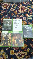 Five Xbox 360 game cases on patterned carpet, four in standard green cases with cover art, one Call of Duty World at War with handwritten insert.