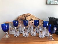 Wide shot showing 6 cobalt blue water goblets with clear barley twist stems and 12 clear etched whiskey glasses grouped on wooden surface.