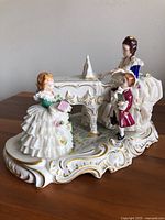 Front and side view of the detailed hand-painted porcelain figurine showing three figures in Dresden lace around a grand piano with gold accents.