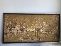 Full view of the large tapestry with a village scene from the 1800s, framed with a dark wood frame and decorative beaded trim.
