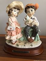 Full front view of the Giuseppe Armani porcelain figurine showing the girl and boy seated, the boy holding a dog, on a wooden base with plaque.