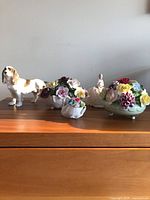 Five porcelain items displayed on wooden surface: porcelain dog on left, three floral bouquets of different sizes, and pincushion figurine with lady figure.