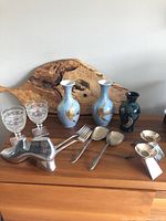 Lot of various items on wooden surface including 2 matching blue lustre vases with gold bird motifs, 1 smaller dark vase, 2 pressed glass wine glasses, white metal shoe bookend, and silver plated serving utensils
