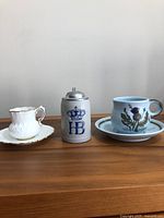 Front view showing the three items side by side on a wooden surface including the white Bone China cup & saucer with gold trim, the German beer stein with pewter lid with blue 'HB' and crown logo, and the light blue Buchanan stoneware cup and saucer with Scottish thistle decoration.