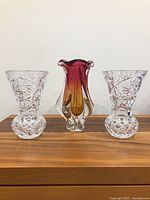 Lot of three Czechoslovakian vases displayed on wooden surface: two clear crystal vases and one pink-orange MCM vase.