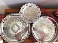 Three silver trays displayed including King’s plate with scrolls and pierced edges, Viking plate with pie crust edges, and medium silver tray with scratches