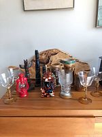 Clear Libbey Tiki Mai Tai glasses with yellow stems and assorted decorative items including totem poles, tiki mugs, and collectible bottles on wooden surface