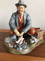 Overall view of Royal Doulton fisherman figurine cooking fish over campfire beside basket and fish on ground.