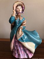 Full view of the Royal Doulton Delphine figurine showing the entire figure in teal coat and pink skirt holding her bonnet.