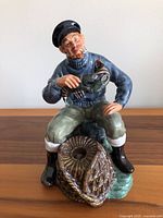 Front view of The Lobster Man figurine showing seated fisherman holding lobster with fishing net in front on wooden surface against white background.