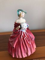 Full view of the Royal Doulton Genevieve figurine showing the red and peach dress and green wide-brimmed hat, holding a white fan with black dots and pink flowers.