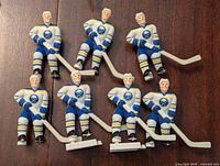 Front view of seven Buffalo Sabres hockey player figurines in blue and white jerseys holding hockey sticks, arranged on a wood surface